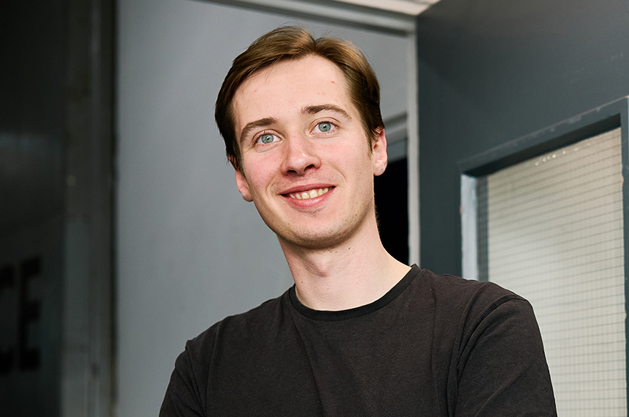 Professional headshot of a team member wearing a dark T-shirt, standing indoors in an office environment.