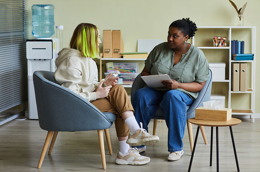 African female social worker talking to teenage girl while they sitting on armchairs at office