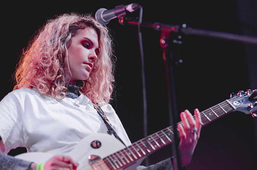 A girl with curly hair playing the guitar with a microphone in front of her.