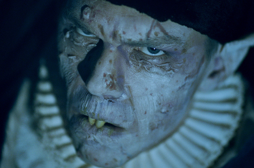 A close-up of a pale, monstrous-looking character with decayed skin, sharp yellowed teeth, and piercing blue eyes.